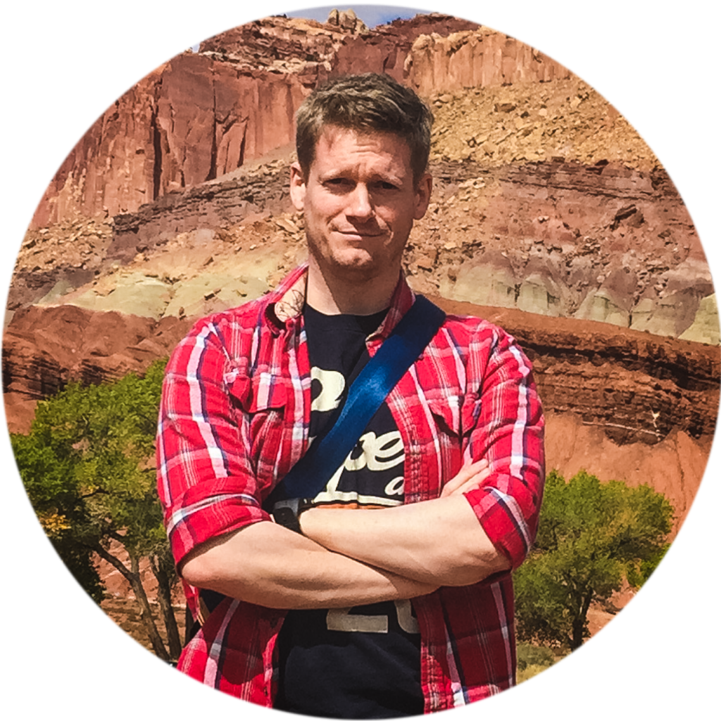 Image of Stephen Cain stood cross-armed in front of tall cliffs of red, orange and yellow rocks.